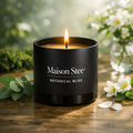Maison Stee Botanical Bliss candle burning in a glass matte‑black vessel on a wooden surface. Fresh greenery, white flowers, and soft natural light create a bright, rejuvenating atmosphere.