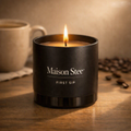 Maison Stee First Sip candle glowing in a glass matte‑black vessel on a wooden surface. Warm café‑inspired lighting with soft shadows evokes a cozy, comforting morning mood.