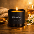 A glass matte‑black Maison Stee candle labeled “Golden Quiet” sits beside an open book and soft florals, lit by warm natural light to evoke a calm, luminous atmosphere.