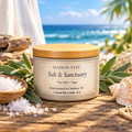 Maison Stee Salt & Sanctuary sea salt and sage scented candle in a cream ceramic vessel with a natural wood lid, surrounded by sage leaves, sea salt, seashells, and driftwood against a serene ocean backdrop.