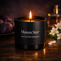 Maison Stee Silk After Midnight candle burning in a glass matte‑black vessel on a dark wooden surface. Warm amber light, soft shadows, and plum and white floral accents create an intimate, luxurious atmosphere.