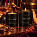 Maison Stee Cognac & Oak candles in glass matte black vessels, lit and glowing, styled beside a cognac glass and raw oak wood pieces on a dark wood surface — Spirit House Collection.
