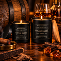 Maison Stee Cognac Barrel candles in  glass vessels beside an aged barrel, cognac glass and decanter, and a resting cigar — Spirit House Collection.