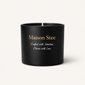 Maison Stee candle in a matte-black vessel with gold branding on a soft cream background