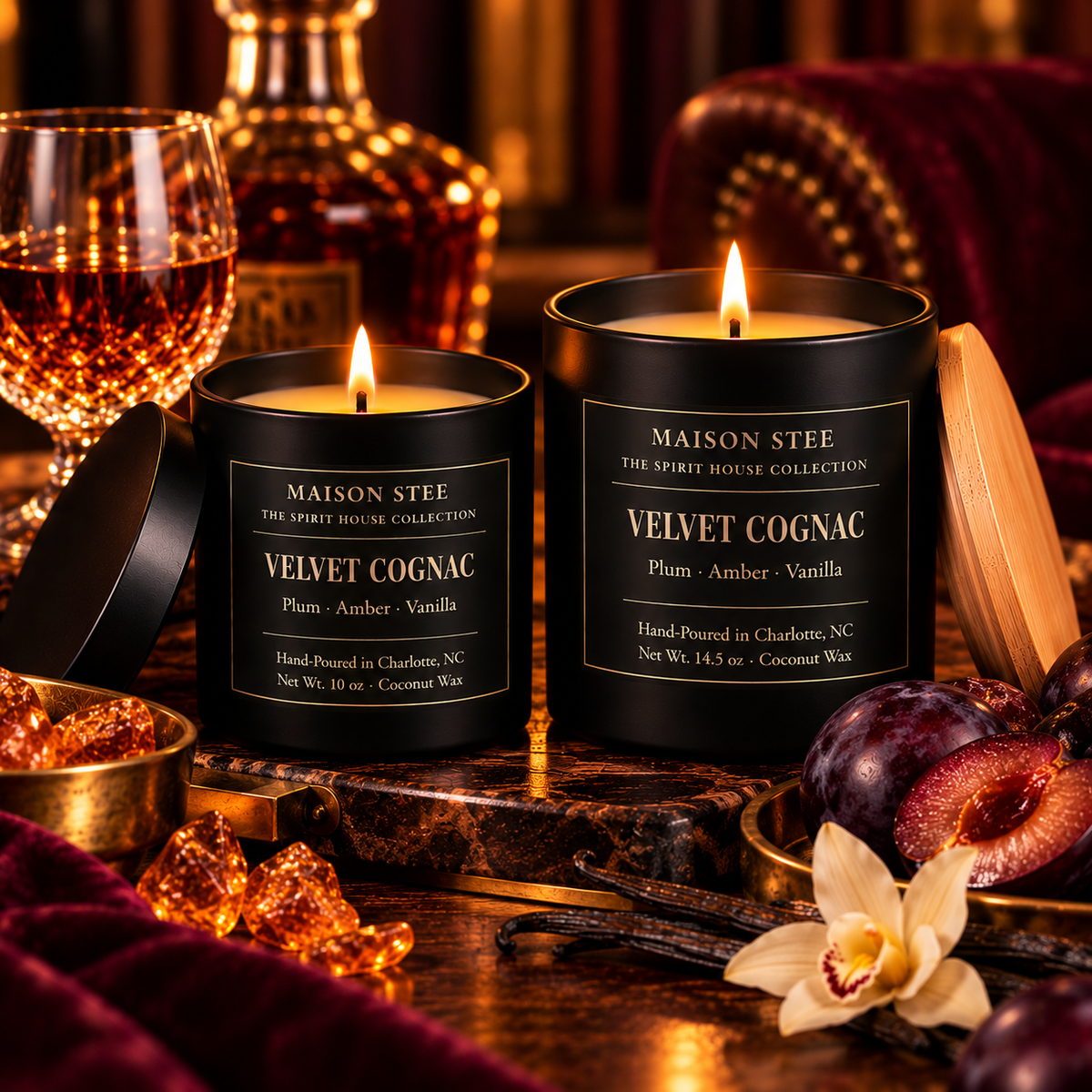 Maison Stee Velvet Cognac candles in glass matte black vessels, lit and glowing, styled beside cognac, rich plums, vanilla, amber accents, and burgundy velvet — Spirit House Collection.